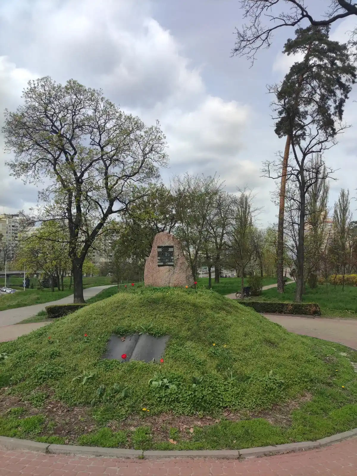 Кам'яний обеліск Василю Стусу на зеленому пагорбі, оточений доріжками, лавами й високими деревами навесні.